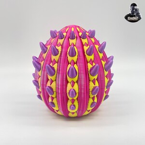 Könnte beinhalten: Eine dekorative, 3D-gedruckte Kugel mit einem auffälligen Farbschema. Die magentafarbene Basis wird durch gelbe und violette Akzente ergänzt. Das Design umfasst vertikale gelbe Streifen und violette, kegelförmige Vorsprünge, die eine strukturierte Optik erzeugen.