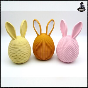 May include: Three decorative Easter bunny figurines in yellow, orange, and pink. The yellow bunny has a textured diamond pattern, the orange bunny has vertical lines, and the pink bunny has a woven texture. Each bunny has long ears.