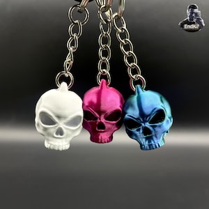 May include: Three skull-shaped keyrings in white, magenta, and blue, each suspended from a silver chain. The skulls have detailed facial features and are 3D printed. The background is a dark grey gradient.