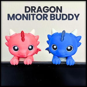 May include: Two 3D-printed dragon figures, one pink and one blue, with white horns and large black and white eyes. The dragons are perched on a black surface, with the text "DRAGON MONITOR BUDDY" above them.