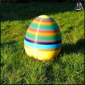 May include: A large, colorful Easter egg with a glossy finish, resting on a bed of green grass. The egg features horizontal stripes in various colors, including yellow, blue, green, orange, and red. The egg is a vibrant decorative item.