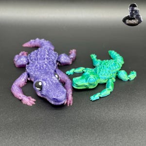 Op de afbeelding: Twee gelede alligatorfiguren. Eén is paars met roze accenten, de andere is groen en blauw. Beide hebben gedetailleerde schubben en witte en zwarte ogen. De figuren liggen op een donkere ondergrond.