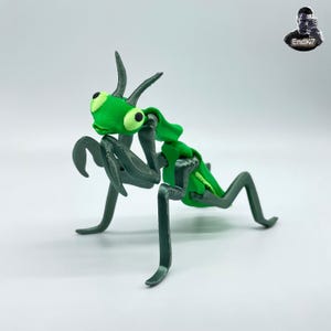 May include: A 3D-printed praying mantis figurine. The insect has a bright green head and body segments, with large, round eyes. Its legs and arms are a darker shade of green. The figurine is posed on a white surface.