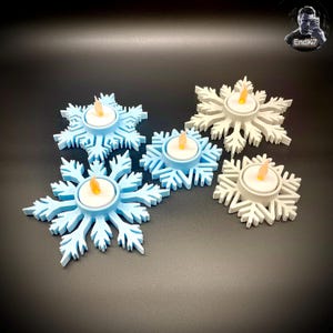 Könnte beinhalten: Schneeförmige Kerzenhalter in Hellblau und Weiß, die jeweils eine kleine, flackernde LED-Kerze halten. Die Halter sind auf einer dunklen Oberfläche angeordnet und schaffen ein winterliches oder festliches Thema. Das Schneeflocken-Design sorgt für eine dekorative Note.