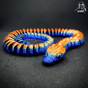 May include: A 3D-printed snake toy in shades of blue and orange. The segmented body curves in a spiral, with the head at the forefront. The snake's scales transition from orange to blue, creating a gradient effect. The toy is on a dark surface.