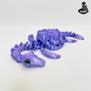 Könnte beinhalten: Eine lavendelfarbene, 3D-gedruckte Skulptur einer Meereskreatur mit einem Blumendesign auf dem Rücken. Die Kreatur hat einen langen, segmentierten Körper, einen Kopf mit einem großen, grünen Auge und vier flossenartigen Anhängseln. Die Skulptur befindet sich auf einer weißen Oberfläche.
