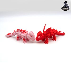 May include: Two dragon figurines, one pink and one red, are positioned facing each other. The pink dragon has red heart accents, while the red dragon has wings. Both have heart-shaped eyes and are on a white background.