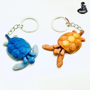 May include: Two turtle keychains. One is blue with gray flippers, the other is orange. Both have silver key rings and chains. The turtles are lying on a white surface.