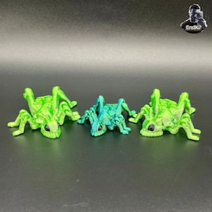 Op de afbeelding: Drie 3D-geprinte insectenbeeldjes in groen- en blauwtinten. De insecten hebben meerdere poten en een gedetailleerd lichaamsontwerp. Elk insect heeft een zwart oog. De beeldjes worden op een donkere ondergrond getoond.