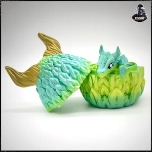 May include: A fantasy-themed sculpture of a dragon emerging from an egg. The egg is two-toned green and yellow, with a textured, leaf-like pattern. The dragon is light blue with large eyes and small horns. The tail is gold and blue.