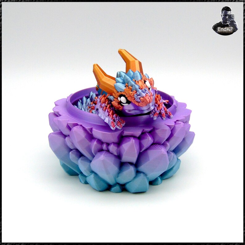 May include: A 3D-printed fantasy creature figurine, featuring a small dragon inside a gem-like container. The dragon has orange horns, blue and red accents, and is nestled in a purple and blue container. The container has a faceted, crystal-like design.