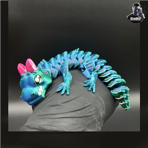 May include: A 3D-printed articulated dragon toy in shades of blue, green, and pink. The dragon has a detailed design with a segmented body, small legs, and a cute face. The toy is flexible and appears to be made of plastic.