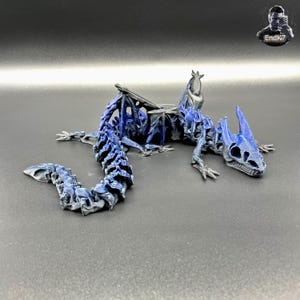Könnte beinhalten: Detaillierte, bewegliche Drachenfigur in Blau- und Grautönen. Der Drache hat ein skelettartiges Aussehen mit komplizierten Details an Körper, Flügeln und Kopf. Die Figur ist auf einer dunklen Oberfläche platziert, um ihr Design zu präsentieren.