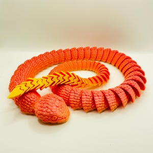 May include: A 3D-printed orange and yellow articulated snake toy. The snake is coiled, with a textured body and a yellow zigzag pattern on its back. Two orange spheres are near the snake's head. The toy is designed to be flexible and poseable.