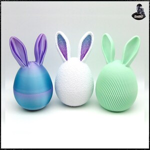 May include: Three decorative Easter bunny egg ornaments. The first is a blue and purple gradient, the second is white with textured surface and blue and purple ears, and the third is mint green with a ribbed design.