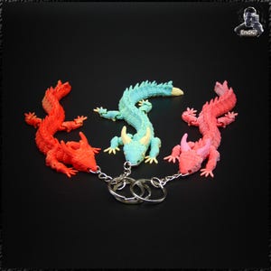 May include: Three colorful dragon keychains in red, teal, and pink. Each dragon has a detailed, textured design and is attached to a silver keyring. The keychains are arranged on a black background.