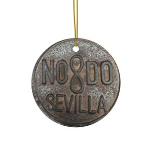May include: A round, rustic, metal ornament with the text "NO8DO SEVILLA" in black lettering. The ornament is hanging from a gold string.