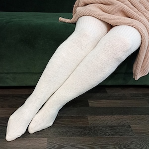 Peut inclure: Chaussettes crème montantes au-dessus du genou. Les chaussettes sont faites d'un tricot épais et texturé. Un pull beige est drapé sur les jambes. L'arrière-plan comprend un canapé vert foncé et un plancher en bois foncé.