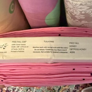 May include: A roll of pink fabric with a label that reads "FREE FALL 108" and "TULA PINK." The fabric is 108 inches wide and made of 100% cotton sateen. The label also includes care instructions and a barcode.