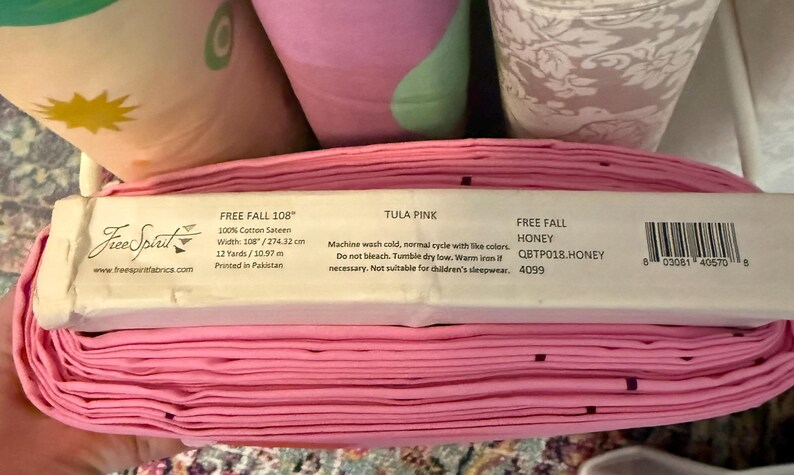 May include: A roll of pink fabric with a label that reads "FREE FALL 108" and "TULA PINK." The fabric is 108 inches wide and made of 100% cotton sateen. The label also includes care instructions and a barcode.