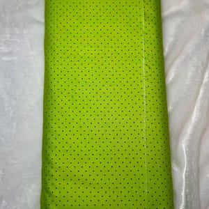 May include: A roll of bright green fabric with a repeating pattern of small, dark blue polka dots. The fabric is tightly rolled and appears to be made of a soft material. The background is a white, textured surface.