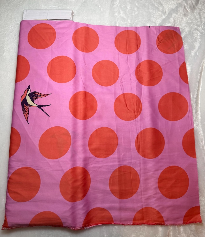 May include: Pink fabric with large orange polka dots and a bird illustration. The fabric is a light pink color with large, evenly spaced orange circles. A bird illustration is on the left side of the fabric.
