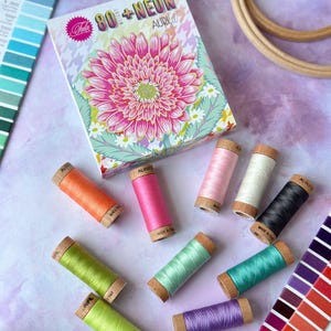 May include: A box of 80+ Neon embroidery floss spools by Aurifil. The box features a pink and white daisy with green leaves. The spools are arranged in a circle around the box. The spools are in various colors, including pink, orange, green, blue, and purple.