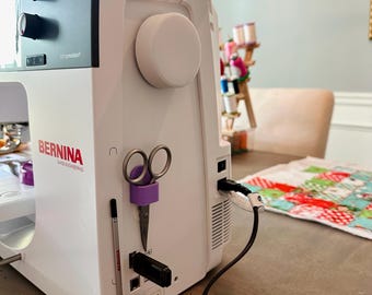 May include: A white sewing machine with a purple magnetic scissor holder attached to the side. The machine has a black power cord and a black foot pedal. The machine is sitting on a wooden table with a colorful fabric on the table.