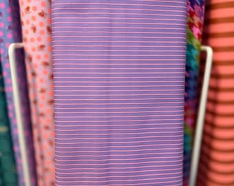 May include: A stack of fabric rolls in a variety of patterns and colors. The primary fabric is a lavender color with thin, horizontal pink stripes. Other fabrics include floral, houndstooth, and striped patterns in pink, green, and orange.