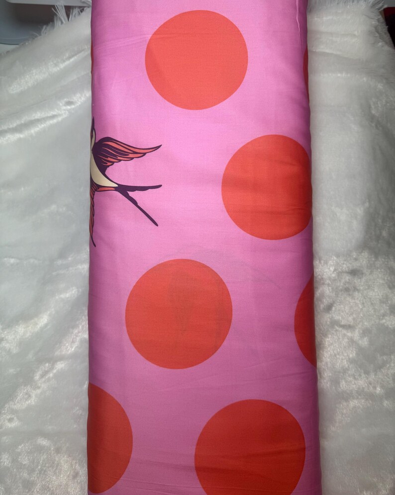 May include: Pink fabric with large orange polka dots and a bird illustration. The fabric is rolled up, showing the pattern of the material. The bird is a stylized design with brown, orange, and purple accents.