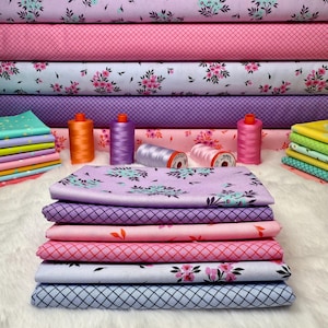 May include: A stack of colorful fabric with floral and geometric patterns. Rolls of fabric in various shades of pink, purple, and blue are stacked behind the folded fabric. Several spools of thread in coordinating colors are also visible.