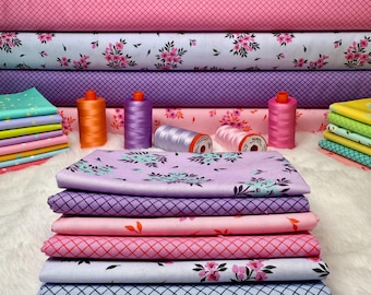 May include: A stack of colorful fabric with floral and geometric patterns. Rolls of fabric in various shades of pink, purple, and blue are stacked behind the folded fabric. Several spools of thread in coordinating colors are also visible.