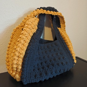 Puede incluir: Un bolso de mano de crochet en color negro y amarillo mostaza. El bolso tiene una base ancha y plana y una parte superior redondeada. Las asas están hechas del mismo hilo que el bolso y son lo suficientemente largas para llevarlas sobre el hombro.