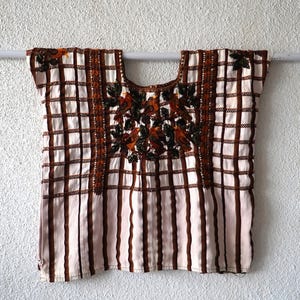 Op de afbeelding: Een blouse met korte mouwen op een crèmekleurige basis met verticale en horizontale bruine strepen. Het borstgedeelte toont gedetailleerd borduurwerk van bruine vogels tussen donkergroene bladeren, omlijst door twee verticale banden met stiksels.