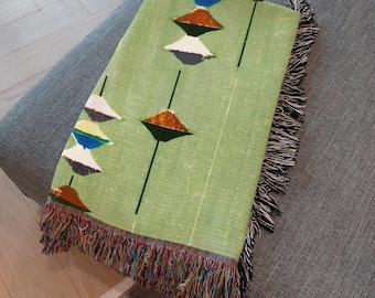 May include: A light green woven throw blanket featuring a repeating pattern of vertical lines and stacked geometric shapes in white, blue, dark green, and orange. The blanket is finished with a multi-colored fringed edge on the bottom.