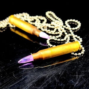 May include: Two bullet-shaped pendants with purple tips and gold-colored casings rest on a reflective black surface. Each pendant is attached to a silver-colored ball chain necklace. The pendants are the focal point, with the chains artfully arranged.