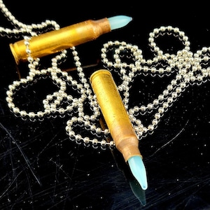 May include: Two bullet-shaped pendants with light blue tips on silver ball chains. The pendants have a gold-colored casing. The chains are arranged in a decorative pattern against a black background.