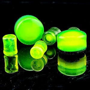 May include: A selection of neon green glass plugs in diverse shapes, including cylinders and rounded forms. The plugs have a translucent quality, with some showing a marbled effect. The objects are arranged on a reflective black surface, creating mirrored reflections.