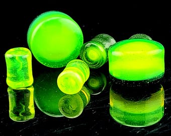 May include: A selection of neon green glass plugs in diverse shapes, including cylinders and rounded forms. The plugs have a translucent quality, with some showing a marbled effect. The objects are arranged on a reflective black surface, creating mirrored reflections.