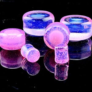 May include: A selection of iridescent glass plugs in various sizes. The plugs display a mix of pink, purple, and blue tones, with a reflective surface. The shapes include rounded and cylindrical designs, suitable for body modification.