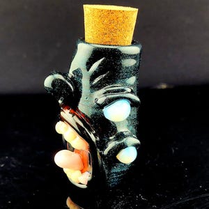 May include: A black glass bottle with a cork stopper. The bottle has a face design with two glowing blue eyes and a pink and yellow mouth. The bottle is likely for decorative use.