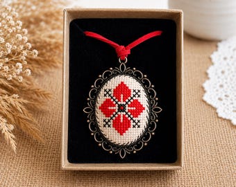 Puede incluir: Un colgante ovalado con un patrón de punto de cruz con motivos florales geométricos rojos y negros sobre un fondo de tela crema, encerrado en un marco de metal oscuro ornamentado y suspendido de un cordón rojo brillante, presentado en una caja marrón.