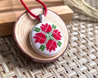 Puede incluir: Colgante redondo con collar de cordón rojo. El colgante presenta un diseño de punto de cruz de flores rojas y hojas verdes sobre fondo blanco, enmarcado en plata. Descansa sobre una rodaja de madera.