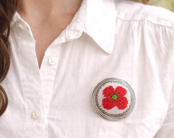 Puede incluir: Broche redondo bordado con una amapola roja y centro verde, enmarcado en un marco de alambre plateado. La broche está prendida a una camisa blanca abotonada, un accesorio de moda.