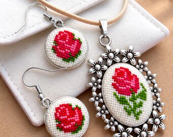 Puede incluir: Un conjunto de joyería que consta de un collar con colgante y pendientes a juego. El colgante es de forma ovalada con un marco floral plateado, con un diseño de rosa en punto de cruz. Los dos pendientes circulares también presentan rosas en punto de cruz.