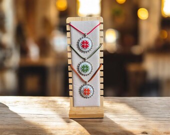 Puede incluir: Tres collares con colgantes bordados en exhibición. Cada colgante presenta un diseño floral en rojo, verde y naranja sobre un fondo blanco. Los collares cuelgan de un expositor de madera con un respaldo de tela beige.