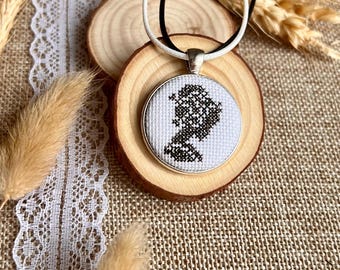 Puede incluir: Un collar con colgante redondo con un diseño de punto de cruz en blanco y negro. El colgante está engastado en un marco plateado y cuelga de un cordón blanco y negro. El collar descansa sobre una rodaja de madera, con elementos decorativos en el fondo.