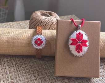 Puede incluir: Un conjunto de joyas hechas a mano que incluye una pulsera y un collar con colgante. La pulsera tiene un diseño redondo bordado con un patrón floral rojo sobre un fondo blanco, sobre una correa de cuero marrón. El colgante tiene un diseño similar.