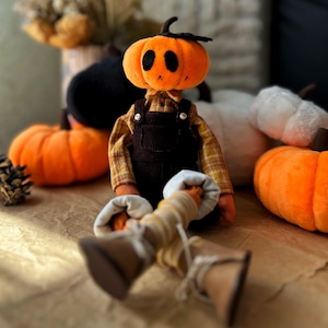 May include: A plush pumpkin-headed doll wearing a plaid shirt and brown overalls. The doll has black eyes and a small black stem. Other plush pumpkins in orange and white are in the background, along with a pinecone.