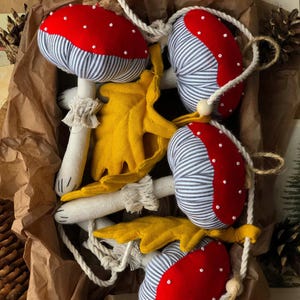 Può includere: Quattro decorazioni di funghi fatte a mano, a strisce rosse e bianche, con foglie di feltro giallo. I funghi sono legati insieme con uno spago bianco.
